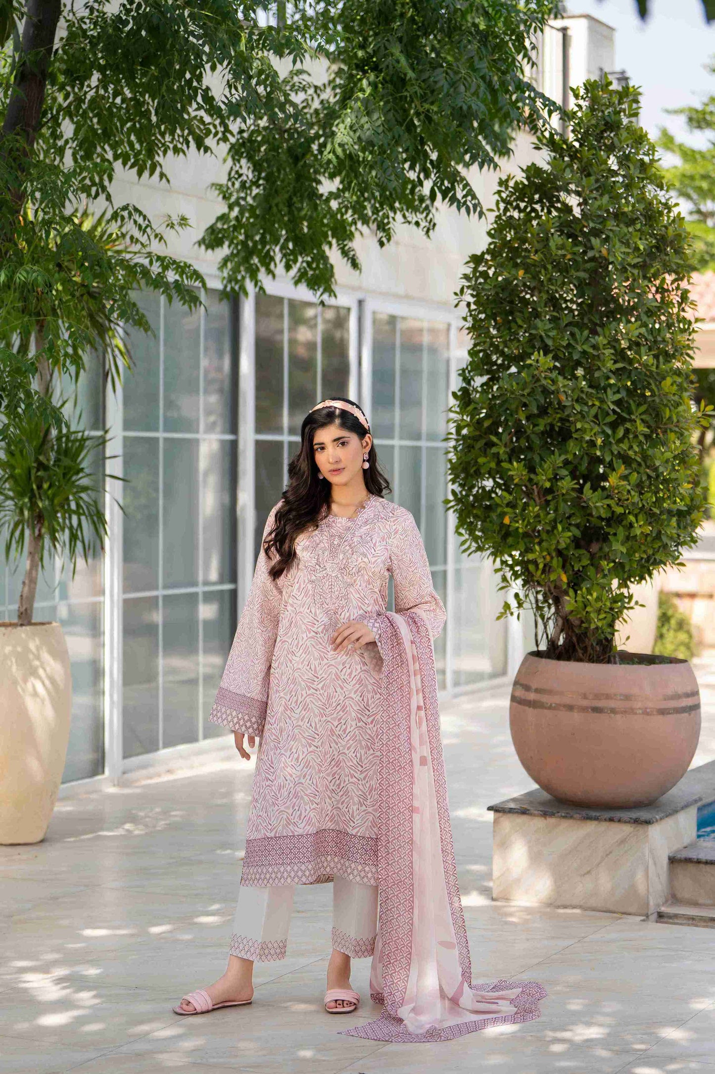 Woman in elegant light pink embroidered suit with white trousers and dupatta, posing outdoors by glass windows and greenery