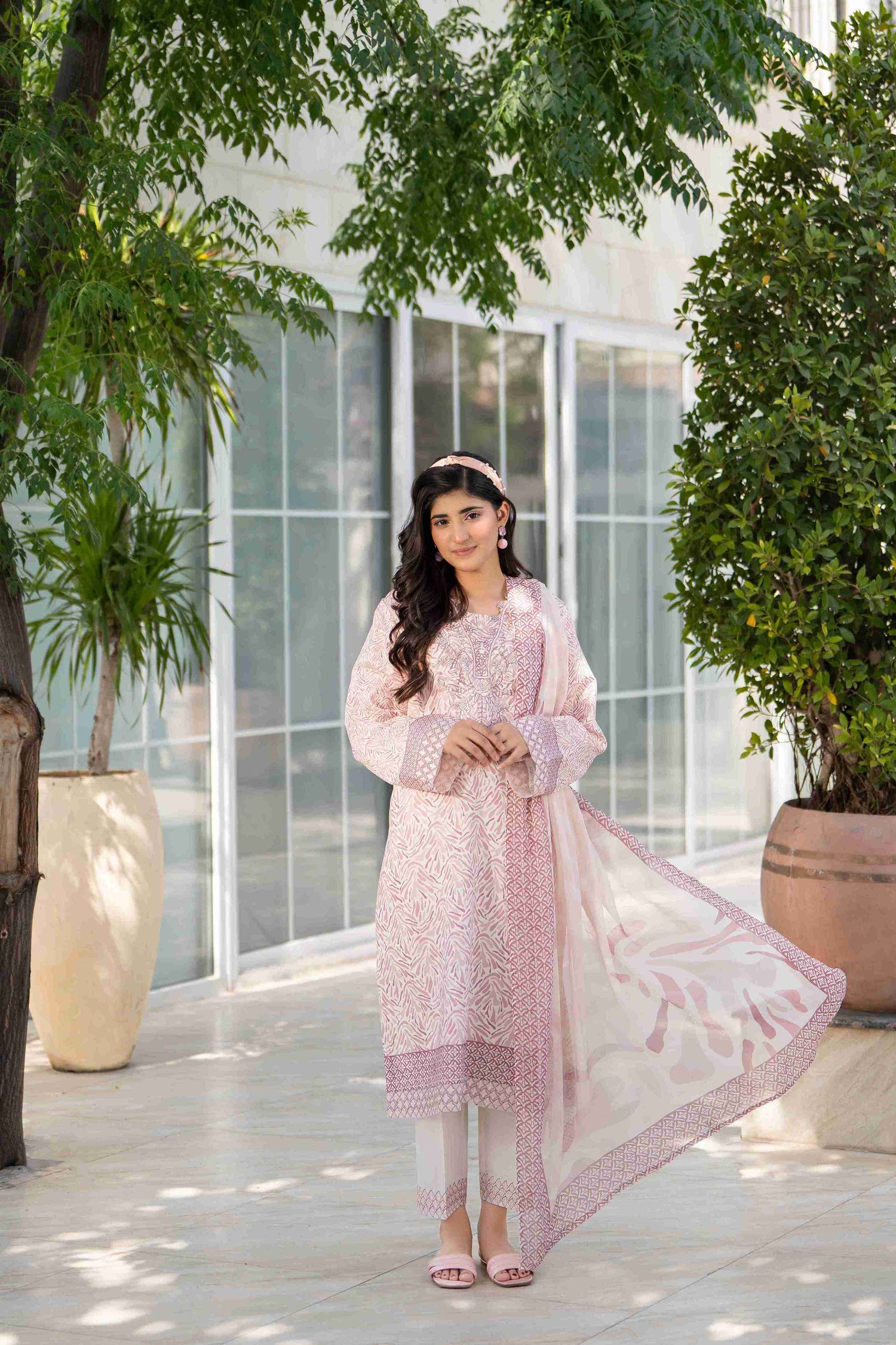 Woman in light pink printed lawn 3-piece suit with embroidered neckline, white trousers, and matching dupatta near greenery