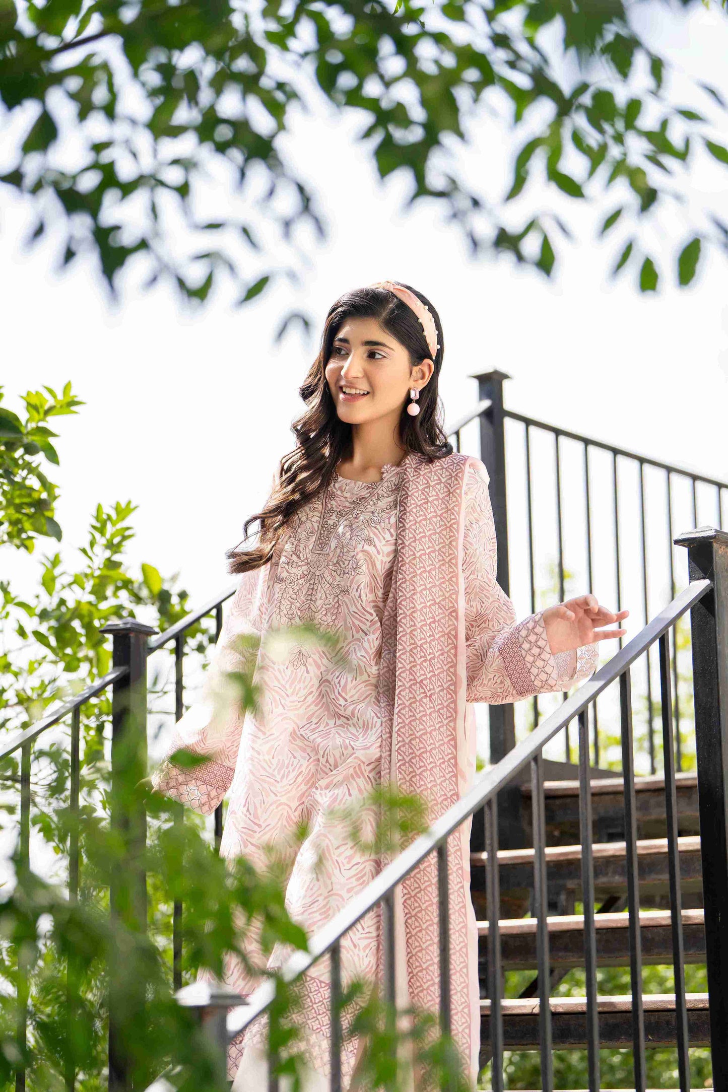 Woman in pastel pink floral lawn 3-piece with ivory trousers and sheer dupatta, walking gracefully in breezy summer style